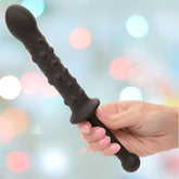 A hand with pink-painted nails holds the CalExotics Rock Bottom Bendies Bendy Beaded Probe, a long black silicone toy with a rounded tip and raised bumps, against a blurred pastel background with circular light bokeh.